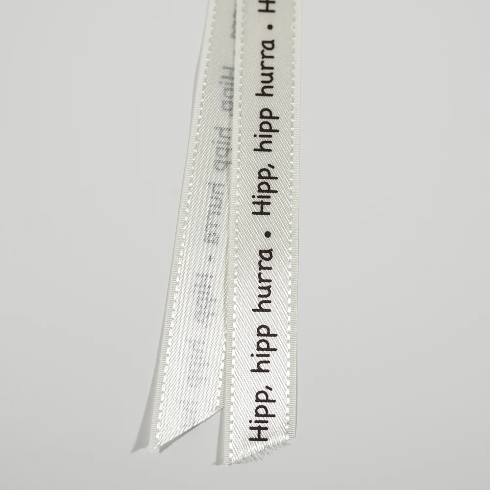 Bånd med tekst kremfarget med brun skrift &raquo; hipp hipp hurra &laquo;. 17mm